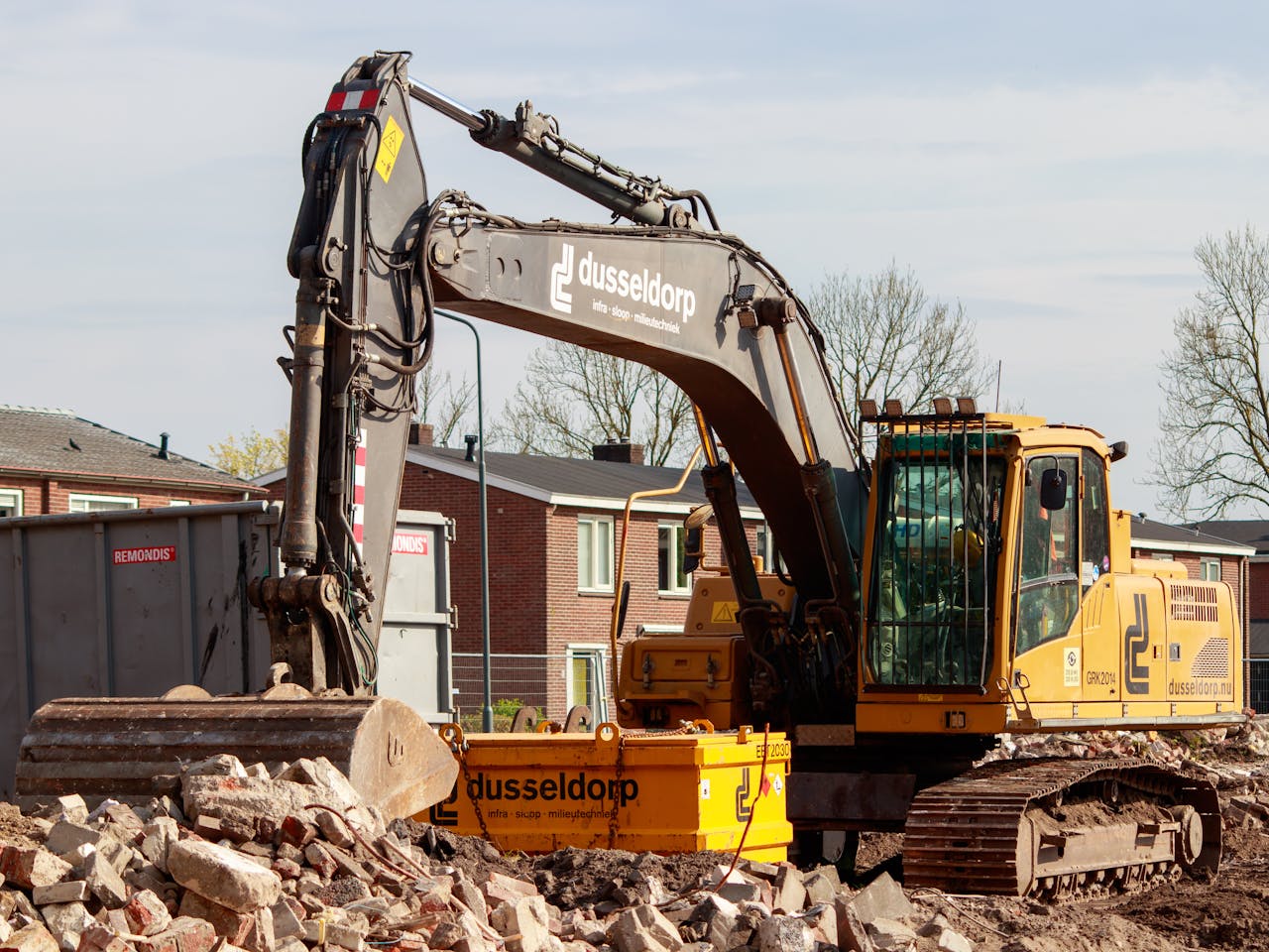 Yellow excavator working on a construction site, surrounded by rubble and buildings in the background.